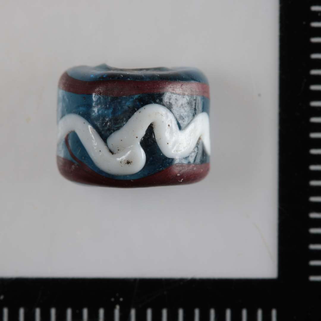 1 glasperle af klart, blåt med indlagte slyng af opakt hvid og rød glasmasse på ydersiden. Halvdelen af perlen er bevaret.