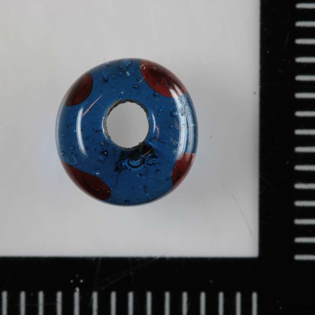 1 perle af fladtrykt kugleform af gennemsigtigt blåt glas med indsmeltede pletter af rødbrunlig uigennemsigtig glasmasse i randen.
