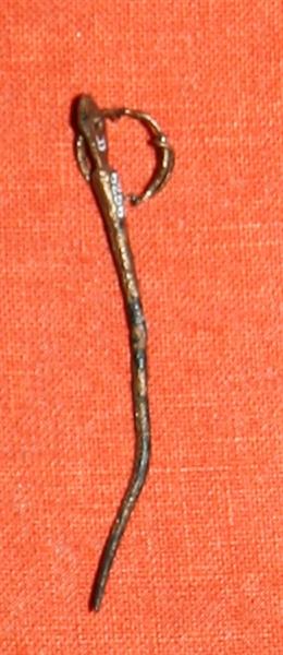 1 ringnål af bronze noget forhøjet henimod den spidse ende. Nålens hoved har flad rhombeagtig form med cirkulært øje, hvor i gennem er trukket en bronzetråd ombøjet i ringform med tynde ender. Længde uden ring : knap 6,4 cm. 