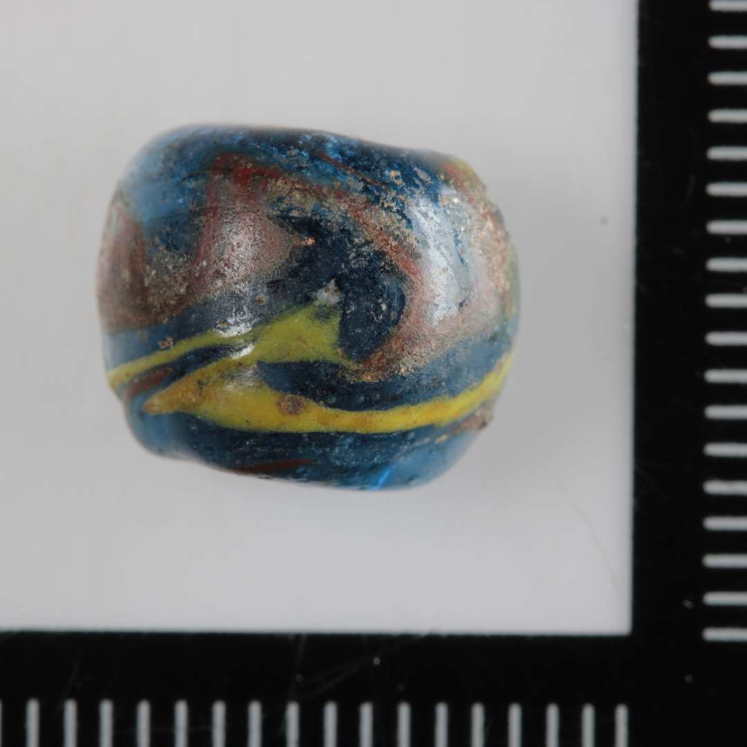 1 perle af blåt glas med pålagt slyng af gult og rødt glas. Halvdelen bevaret.