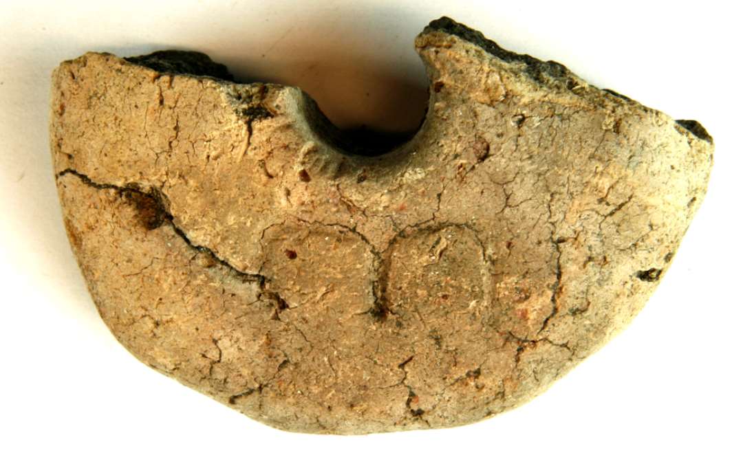 1 fragmenteret vævevægt af brændt ler med indstemplet ornamentik eller bomærke på oversiden. Diameter: 9,5 cm.