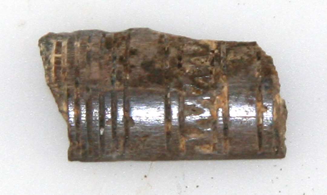 1 fragmentstump af fladhvælvet kamskinne af hjortetak. På oversiden er stykket forsynet med en ornamentik af tværgående stregfurer samt et enkelt bælte med en art zig-zag bånd. L : 1,8 cm. Br : 1,0 cm.