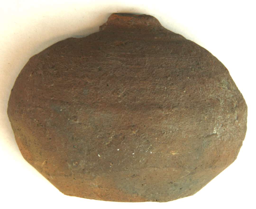 Halvdelen af sparebøsse af rødbrændt lergods af fladtrykt kugleform med plan standflade og lille flad topformig afslutning foroven. Bunddiameter: 5 cm., højde: 6,8 cm.