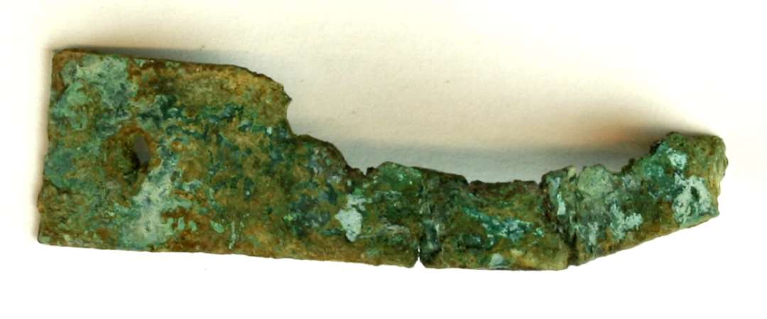 1 fragmenteret tynd ombøjet bronzestrimmel med et indslået hul i den ene ende. Bredde godt 1 cm. Længde i ombøjet stand, ca. 3,5 cm.