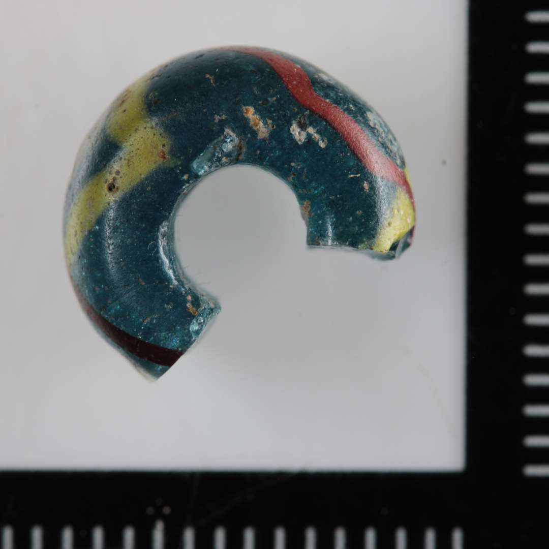1 fragmenteret skiveformig afrundet perle af gennemsigtigt blåligt glas med korsvis indlagte gule og rødbrune tråde i kanten afvekslende med cirkulære indlægninger af hvidt glas.