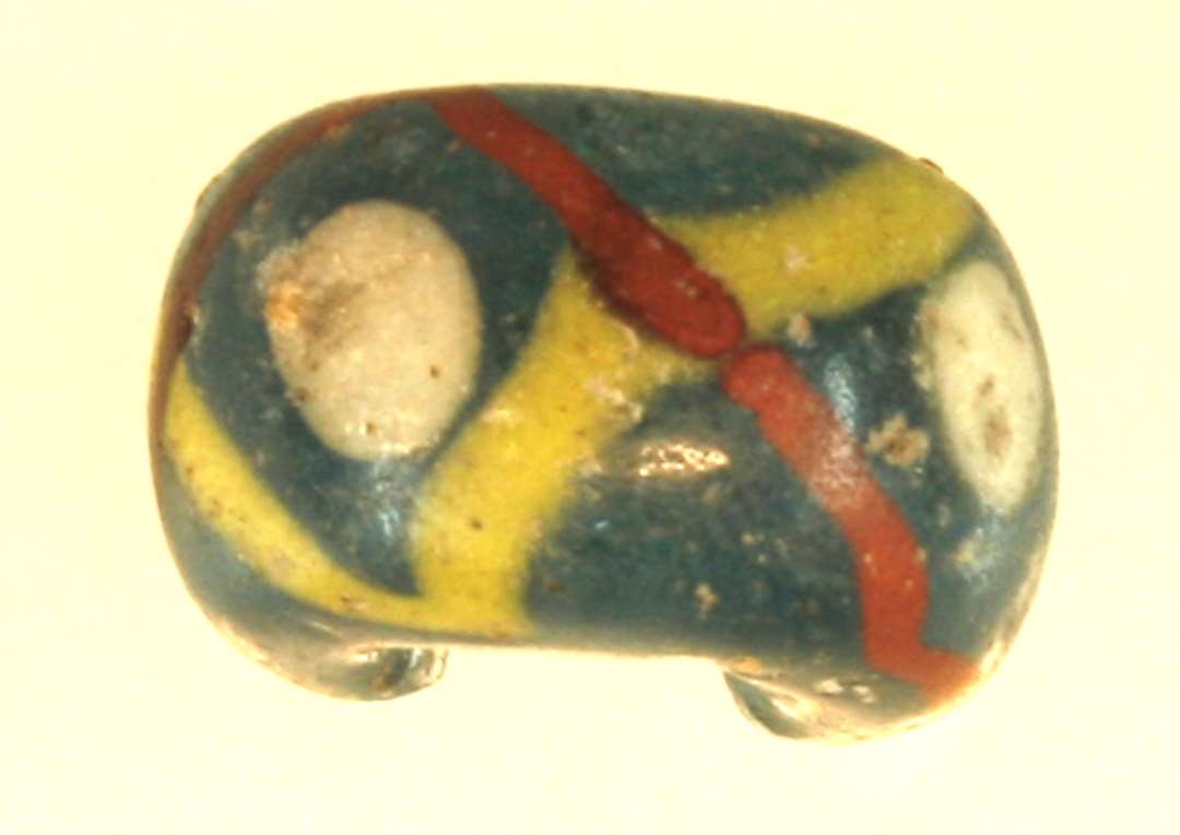 1 fragmenteret skiveformig afrundet perle af gennemsigtigt blåligt glas med korsvis indlagte gule og rødbrune tråde i kanten afvekslende med cirkulære indlægninger af hvidt glas.