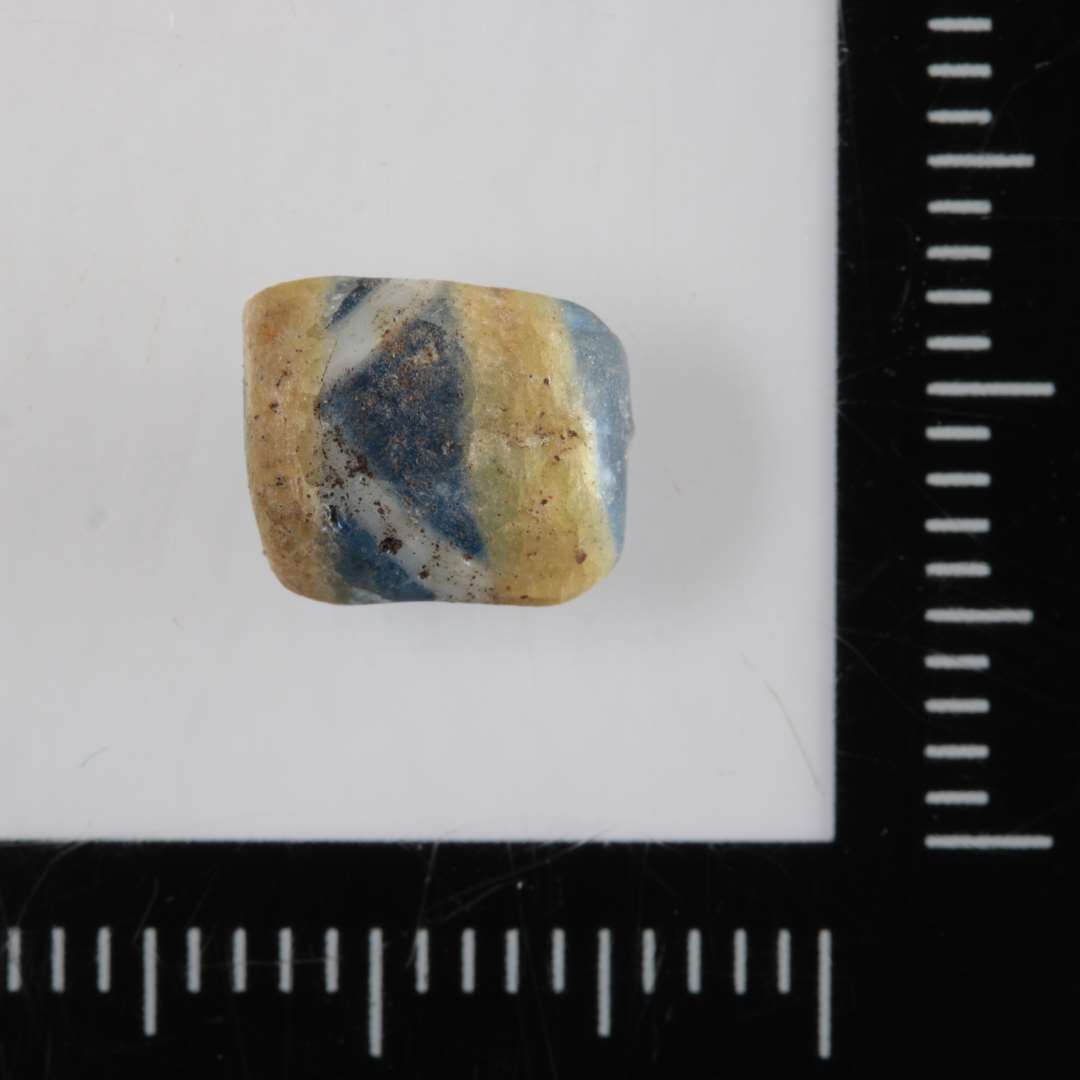 1 fragment af cylinderformet perle af delvis gennemsigtigt blåligt glas med indsmeltede gule kanter, hvorimellem hvide streger i en art zig - zag - forløb.