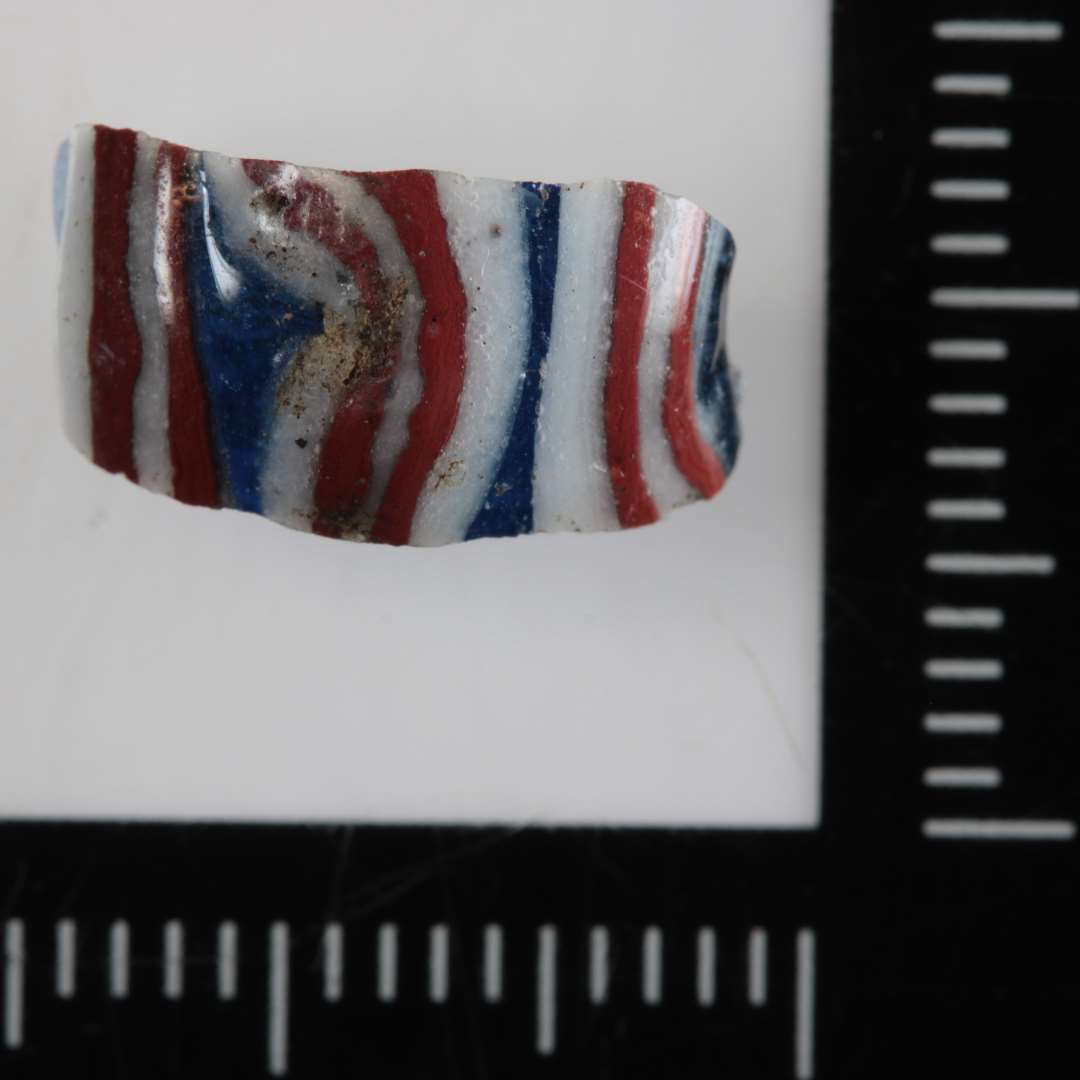 1 fragment af afrundet konisk ( tøndefomet ) perle af blåligt gennemsigtigt glas med indstemlede rød- og hvidstribede streger og bølgelinier af uigennemsigtig glasmasse på ydersiden. h: 13 mm.