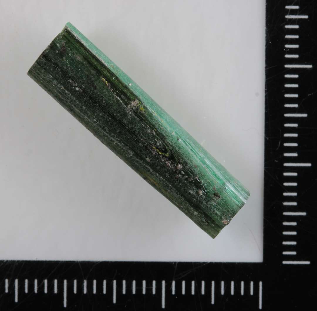 1 stump råglas af grønlig, delvis gennemsigtig farve. Stykket har aflang stangform med svagt rillet overflade og rektangulært tværsnit. L : 2,55 cm. Tværmål : 0,45 x 0,7 cm.