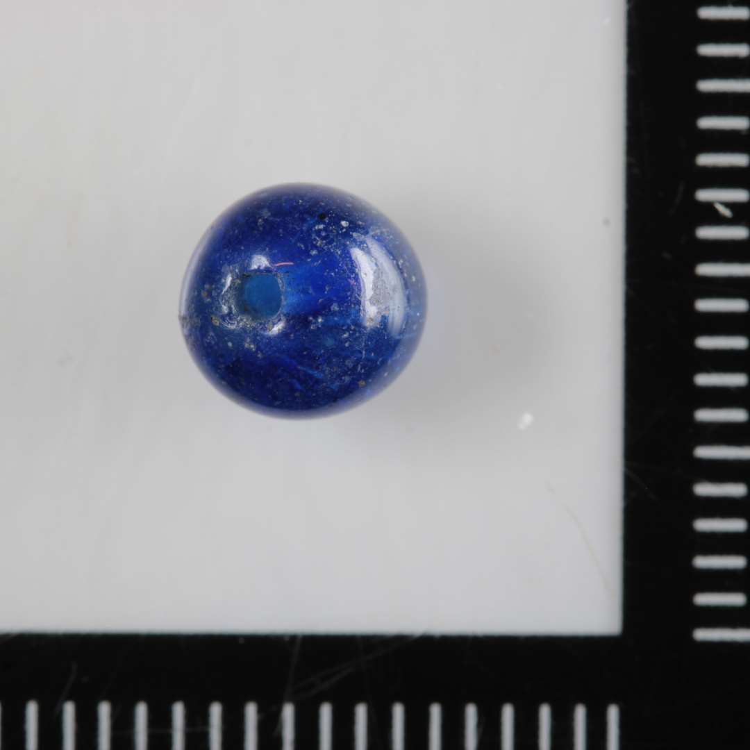 1 omtrent kugleformet perle, let fladtrykt i den ene ende af dybblåt gennemsigtig glas. 6 mm.