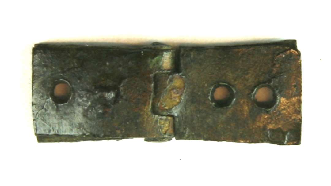 Miniaturehængsel betegnet EO af bronze eller messing tildannet af to ombøjede rektangulære pladestykker, hvis ombøjede ender er indpasset i hinanden i et hængselled sammenholdt med en gennemtrukket bronzenitte. Hver hængselhalvdel er forsynet med to gennemboringer, og i en af disse sidder en søm eller naglestump fastrustet. St. L. 2,9 cm.  St. br. 1,1 cm.