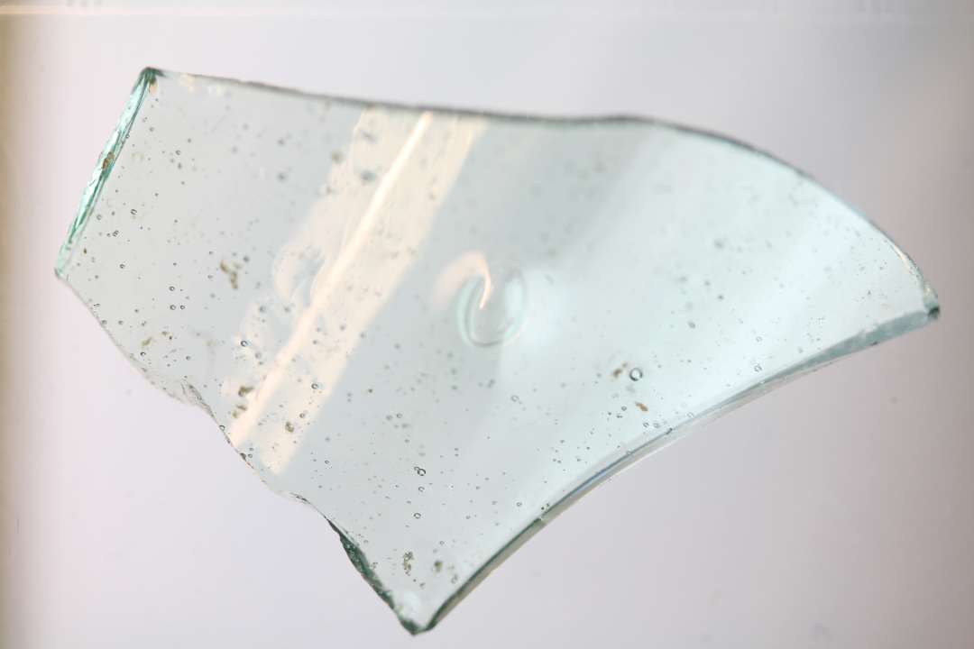 1 krumt sideskår fra bæger af svagt grønligt gennemsigtigt glas hvori en større og mange mindre, fine blærer. Længde: 2,9 cm. Bredde: 2,0 cm. vægtykkelse: ca 12 mm.