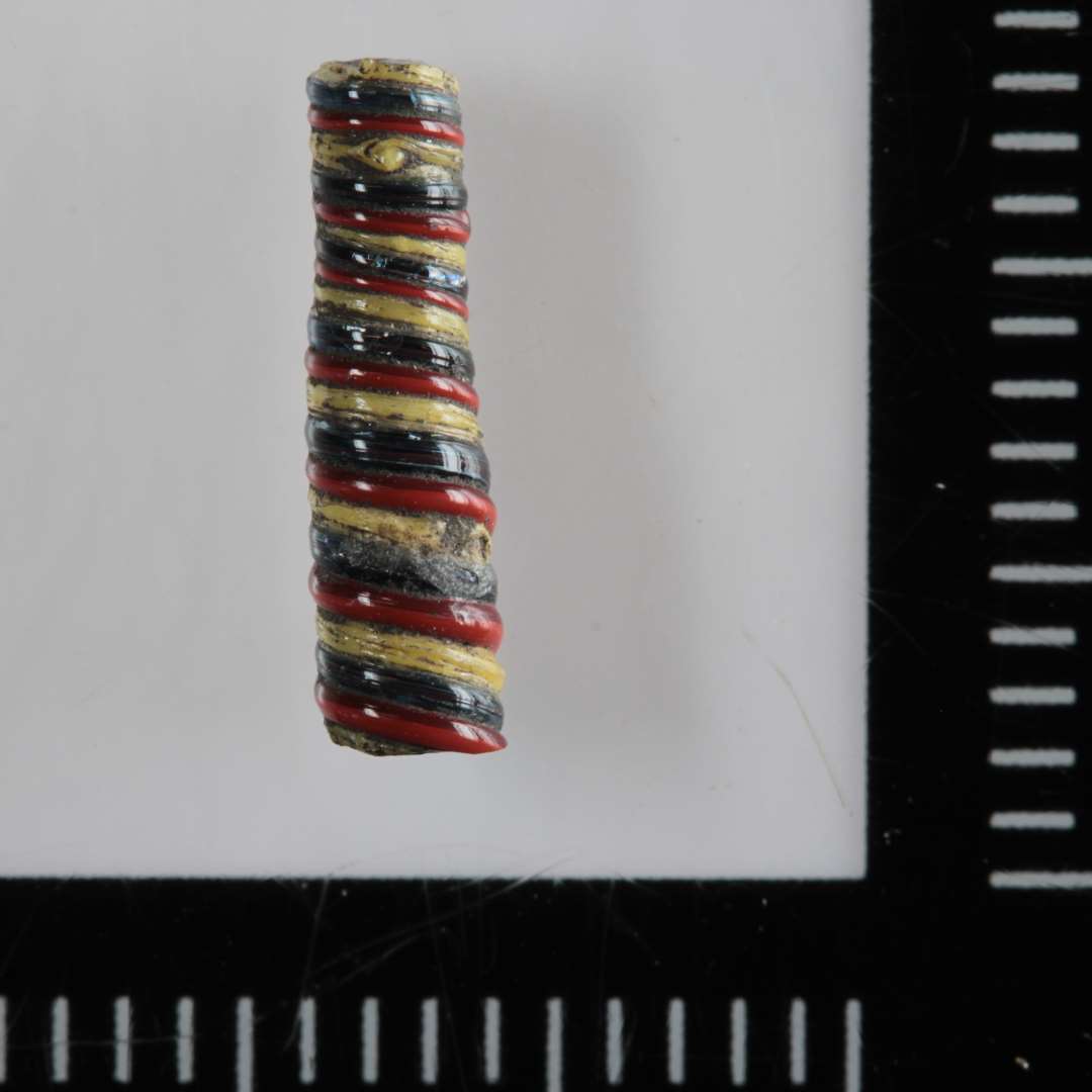 1 fragment af let konisk glasstang af blåligt gennemsigtigt glas med bevikling på ydersiden af tynde tråde af gult og rødbrunt ugennemsigtigt glas. Diameter: 0,3 cm. Længde:   1,1 cm.