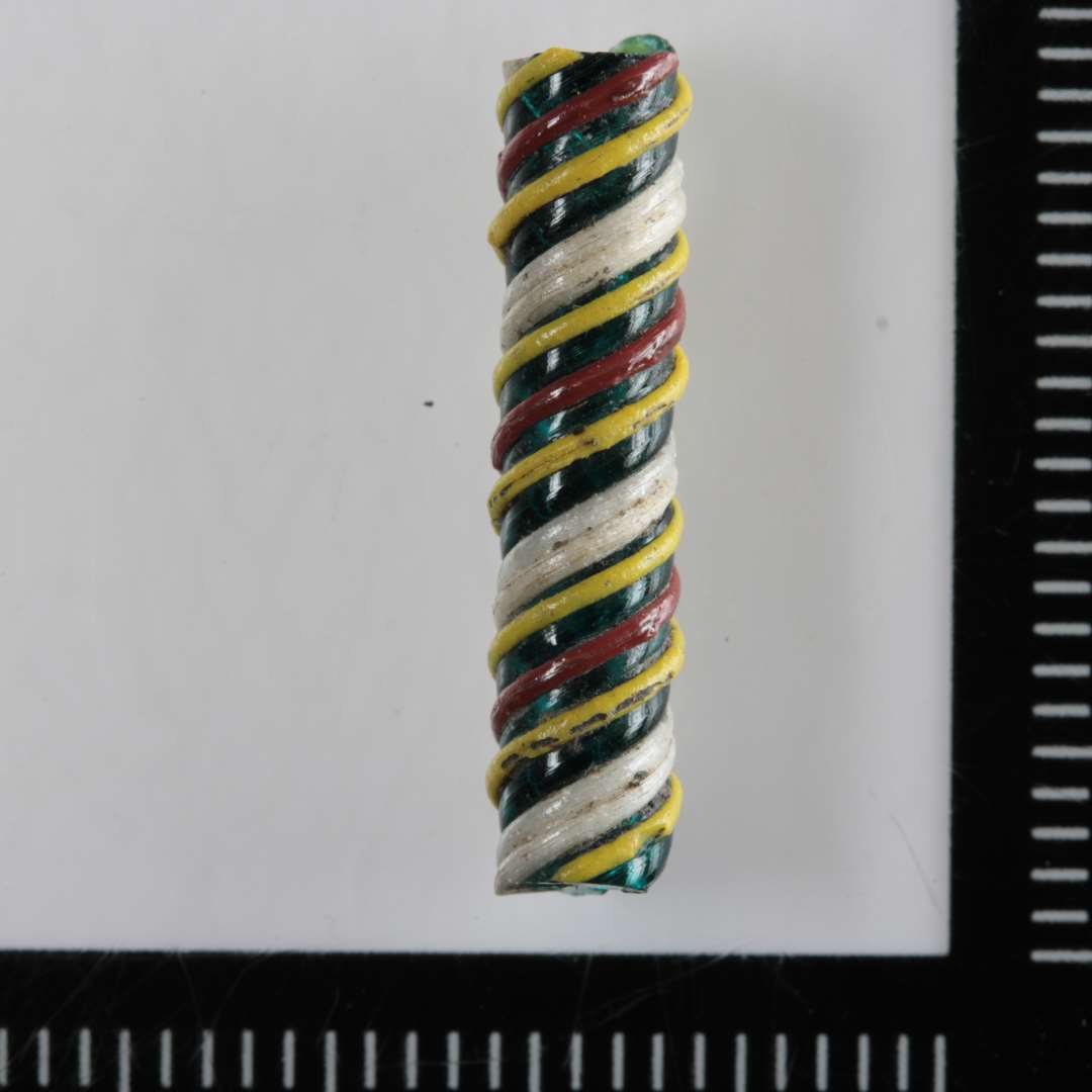 1 fragment af cylindrisk glasstang af blågrønlig gennemsigtig glasmasse, hvis yderside næsten  er dækket af omsnoede tynde tråde i z-snoet forløb af hvidt, gult og rødbrunt, ugennemsigtigt glas. Længde:   2,0 cm. Diameter: 0,4 cm.