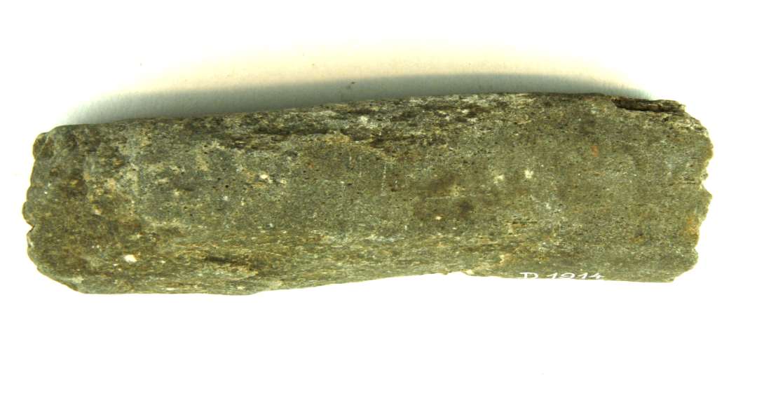 Fragment af stangformet hvæssesten af lysegrå sandsten. Fragmentet udviser en skæv og uregelmæssig, firsidet tværsnitsform med stærk nedslidning henimod den ene ende, som er overbrudt. Den modsatte oprindelige ende har tyk og klumpagtig form. Største længde: 13,15 cm. Bredde: 3,5 cm. Tykkelse: 3,5 cm.