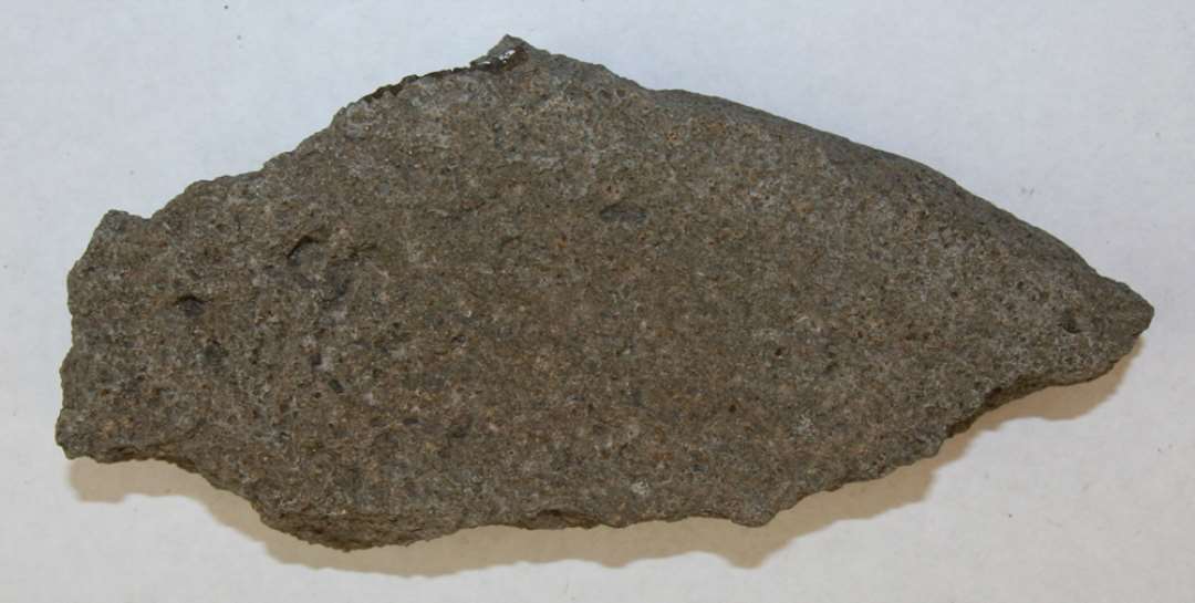 Fragment af overdel af kværn af basalt. Mål: 16x7 cm. Ca. 3 cm tyk. Ret ujævn overside 'grube'-hugget. Del af dreje'-hul bevaret.