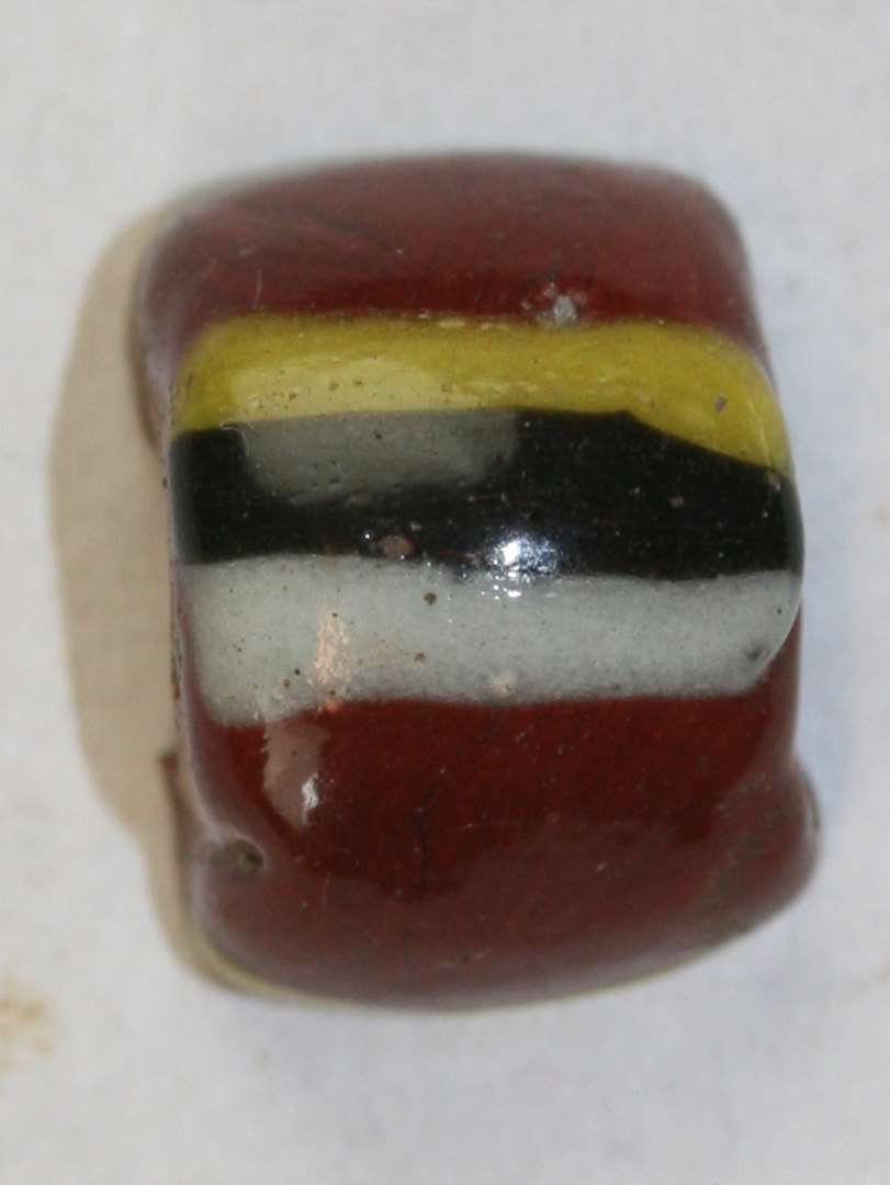 Halvdelen af cylindrisk perle. Rød, uigennemsigtig med gul, sort og hvid tværgående stribe. Største mål: 1,3 cm.