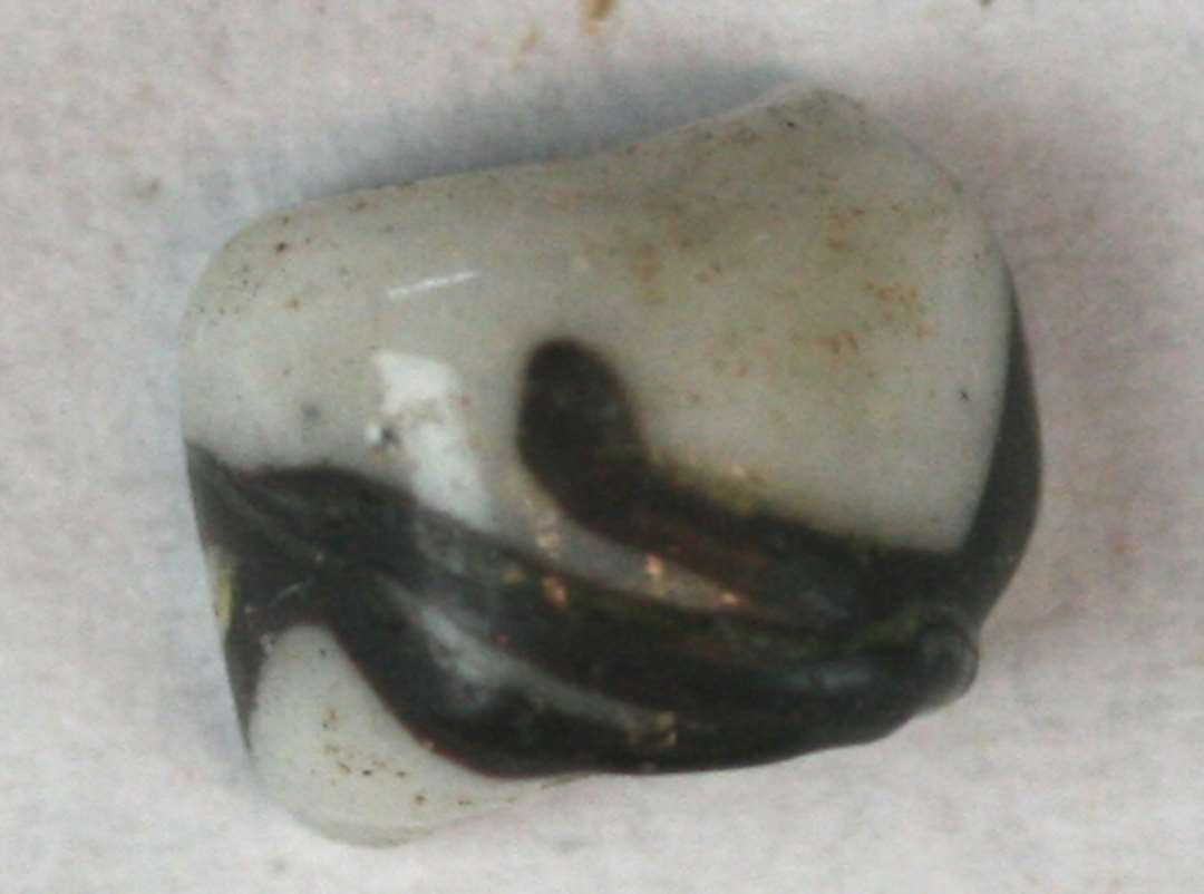 Halvdelen af cylindrisk, uigennemsigtig hvid-grå perle med sort dekor i form af slyng. Største mål: 0,8 cm.