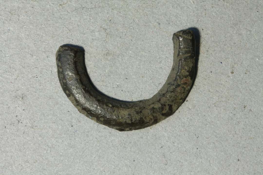Halvdelen af et lille ringspænde, hvælvet overside med rester af tværstreger (?), flad bagside. Diam: 21 mm.