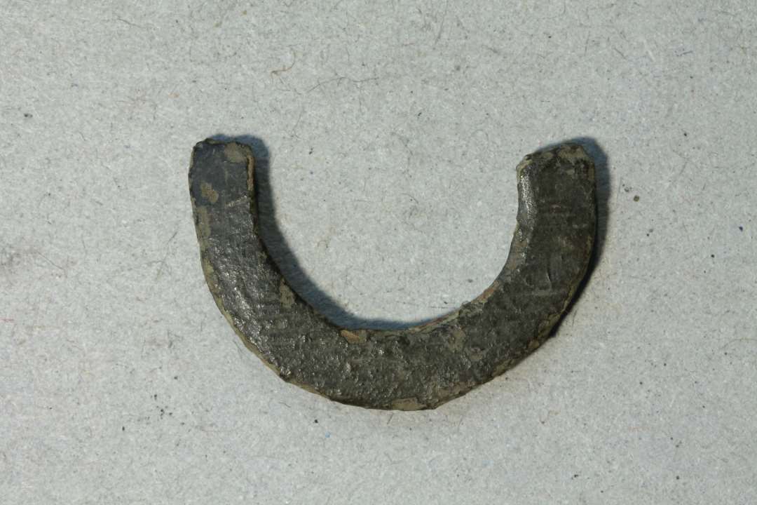 Halvdelen af et lille ringspænde, hvælvet overside med rester af tværstreger (?), flad bagside. Diam: 21 mm.