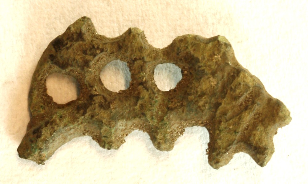 Fragment af fibel i gennembrudt arbejde af kobberlegering med rest af nålefæste på bagsiden. Største mål: 22 mm.