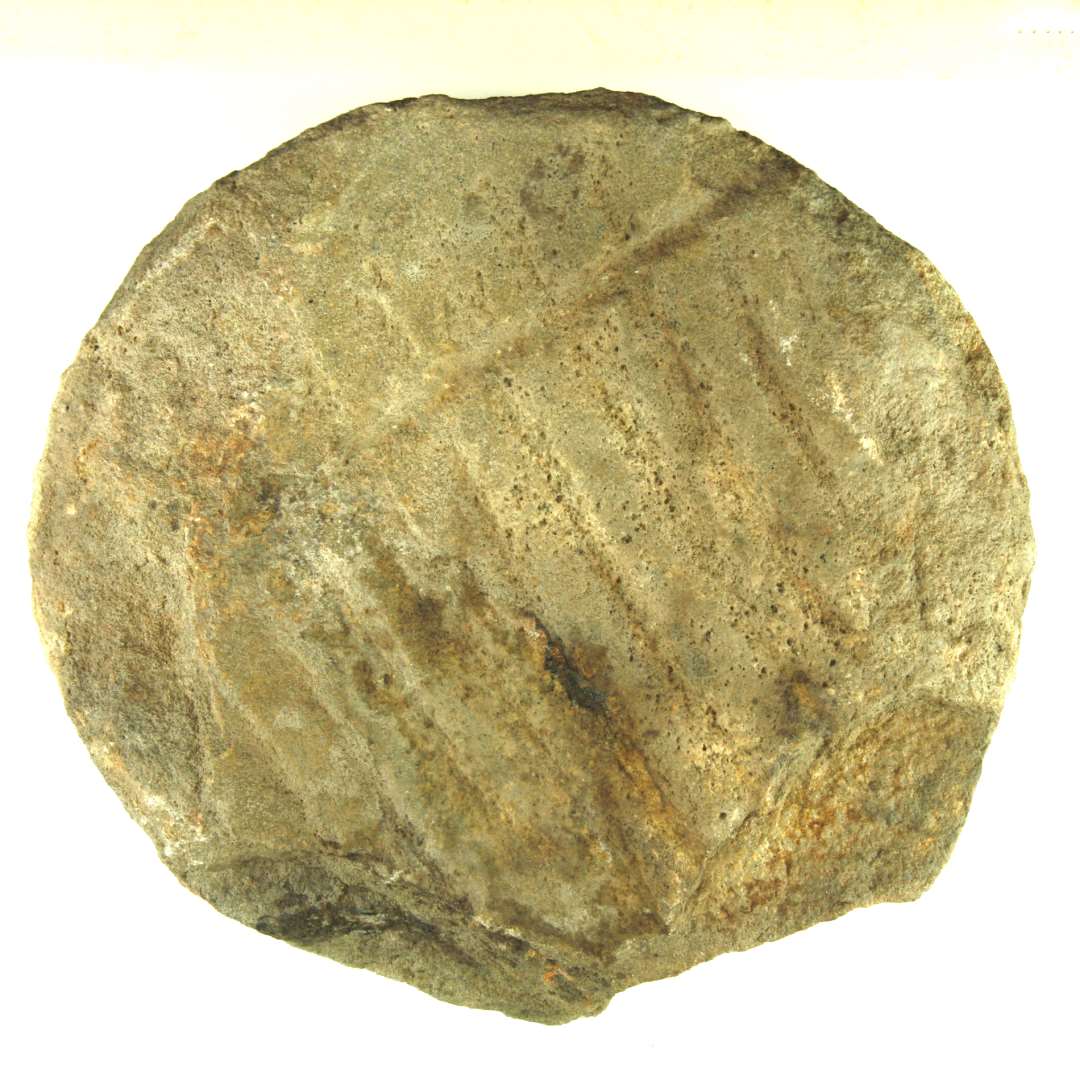 Fragment af kværnsten af finkornet, grålig granit. Cirkulær form med riflet overside. Sekundært tildannet og anvendt som sokkelsten i huset under bindingsværkssøjlen i sekundær skillemur.  Mål: 25 cm. Tykkelse: 9 cm.