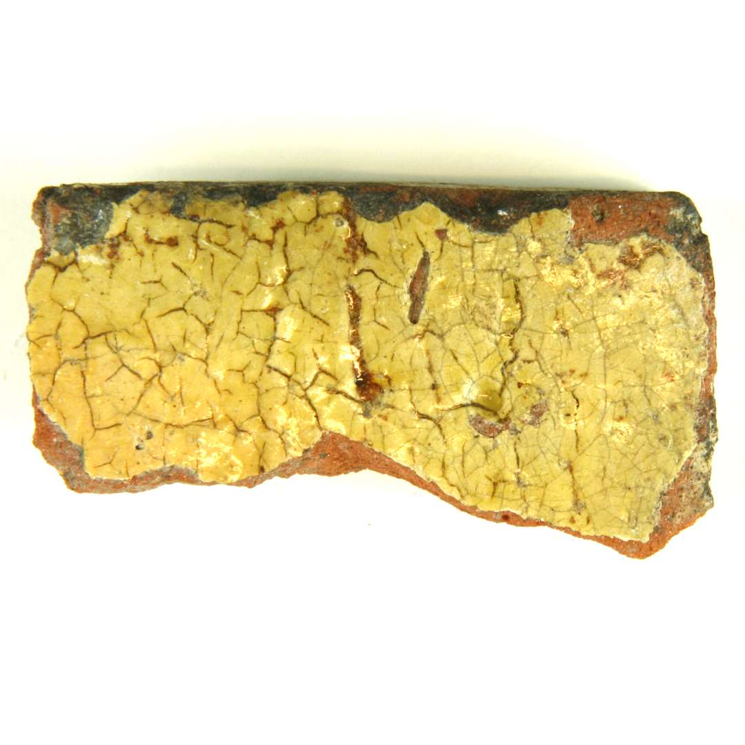 Fragment af gulvflise af rødbrændt ler. Overfladen begittet og klart blyglaseret.  Mål: 6,5 x 3,5 cm.