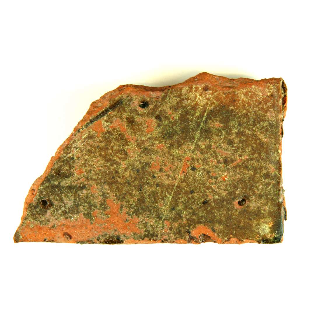 Fragment af gulvflise. Overfladen glaseret med grønt blyglasur, nedslidt. Mål: 11,5 x 7,0 cm. Tykkelse: 2,2 cm.