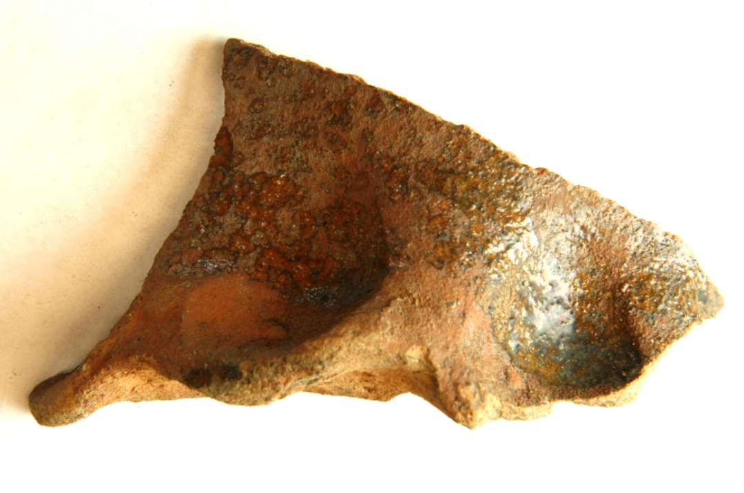 1 bund/sideskår fra større kande af rødbrændt lergods med fremtrædende fodrand med kraftige fingerindtryk. Kantsiden er delvis dækket af ujævn plettet klar blyglasur.
