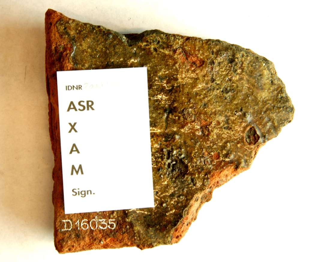 1 hjørnefragment af gulvflise af rødbrændt lermasse med ujævn klar til grønlig blyglasur på oversiden. Største mål: 12,7 x 12,9 x ca. 3 cm.