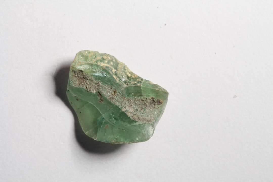 Fragment af polyhydrid, polyedrisk glasperle. Halv-gennemsigtig, grøn. Overfladen nedbrudt. Diameter: ca 1 cm.