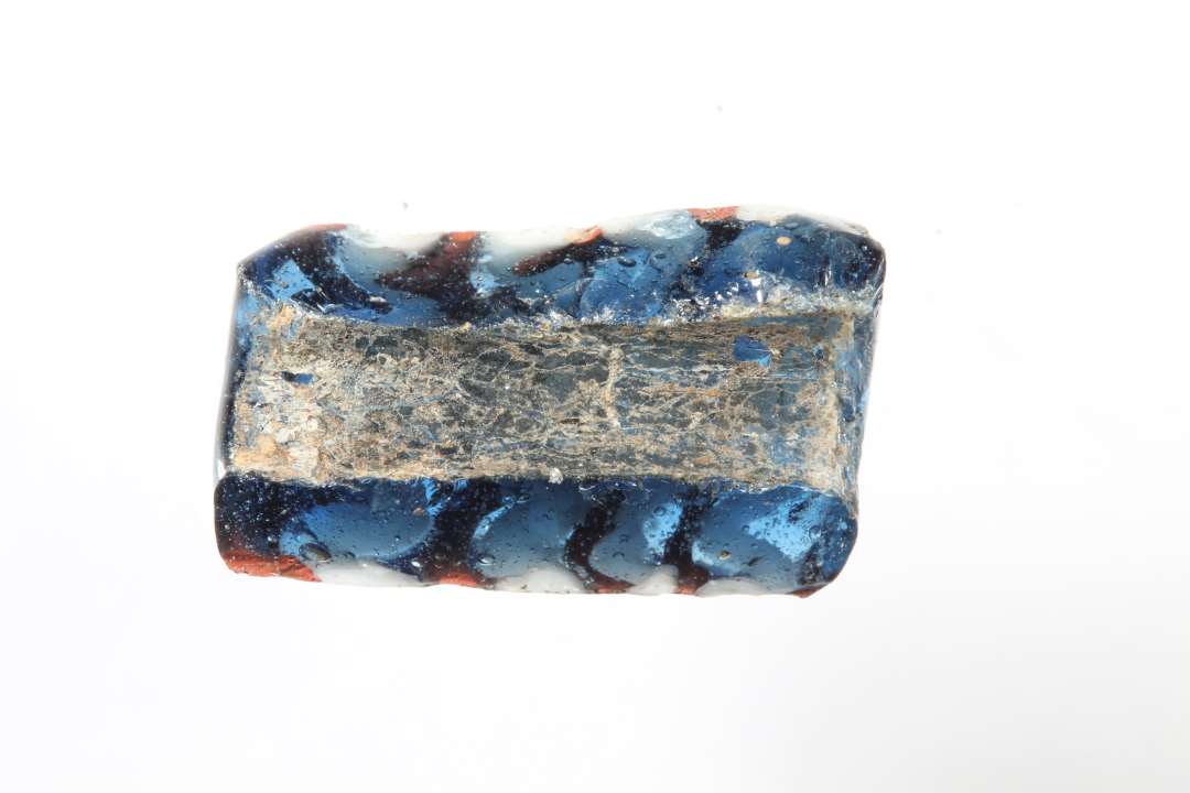 Fragment af cylindrisk ribeperle, gennemsigtig, blå med hvidt og rødt. Største mål: 1,5 cm.