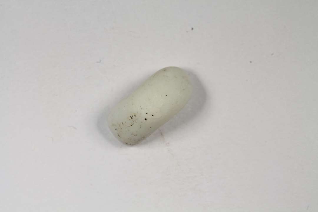 Halv cylindrisk eller ringformet, uigennemsiigtig, hvid-grå glasperle. Diameter: 1 cm.