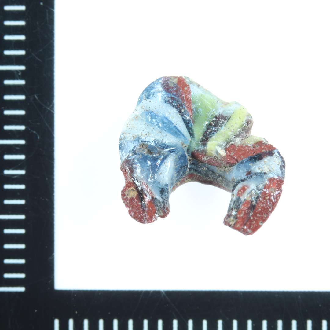 Fragment af stor mosaikperle. Gennemsigtig blå med rødbrunt, gult  og hvidt. Største mål: 1,5 cm.