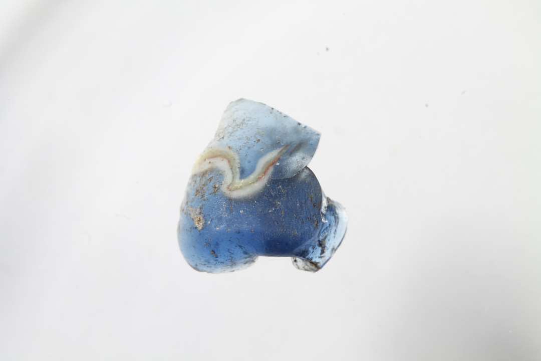 Fragment af cylindrisk kubisk ribeperle. Gennemsiigtig blå med rød og hvid dekoration. Slidt overflade. Største mål: 1,1 cm.