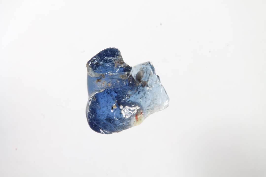 Fragment af cylindrisk kubisk ribeperle. Gennemsiigtig blå med rød og hvid dekoration. Slidt overflade. Største mål: 1,1 cm.