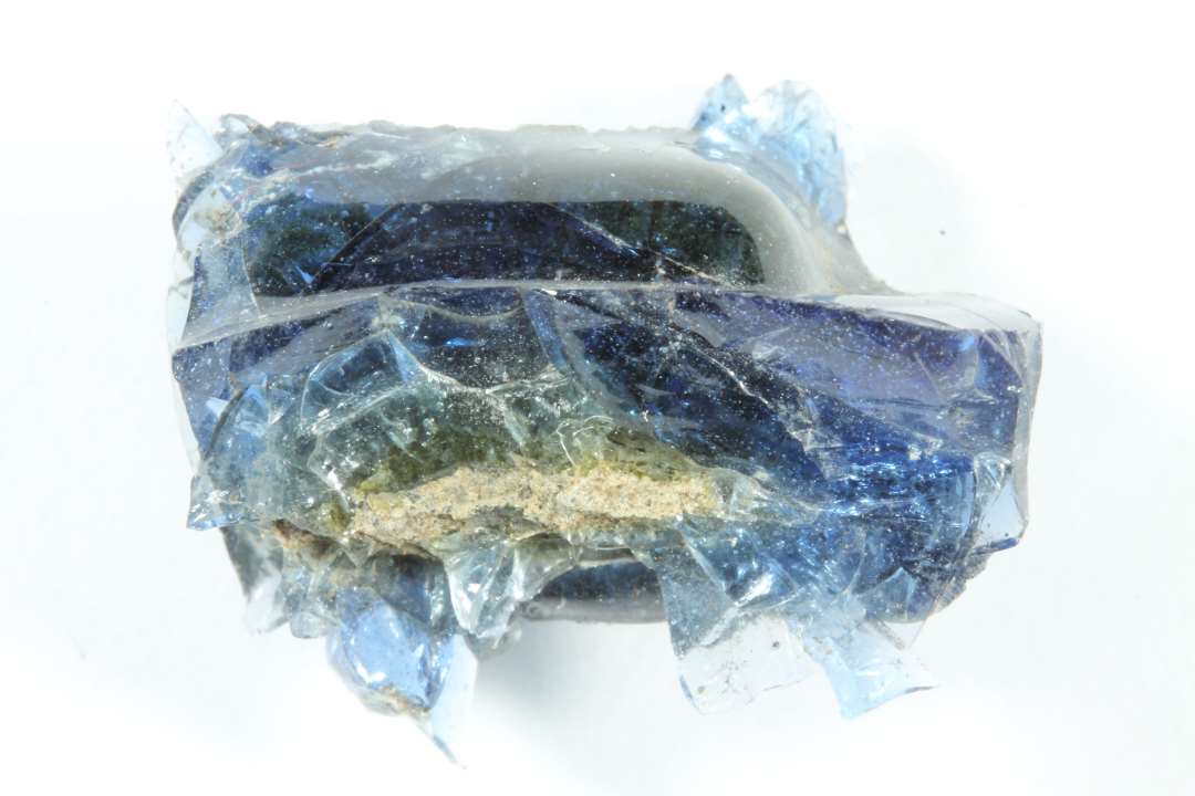 Råglas, gennemsigtig blå. Fastsmeltet til en rektangulær blok af ler (?) eller sten (0,8x0,8x1,2 cm). 