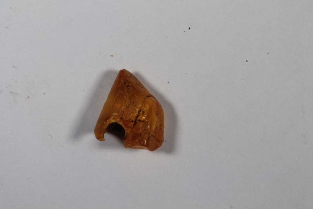 Fragment af ravperle. Knækket i gennemboringen, Uregelmæssig facon/grov forarbejdning. 1x0,7 cm.