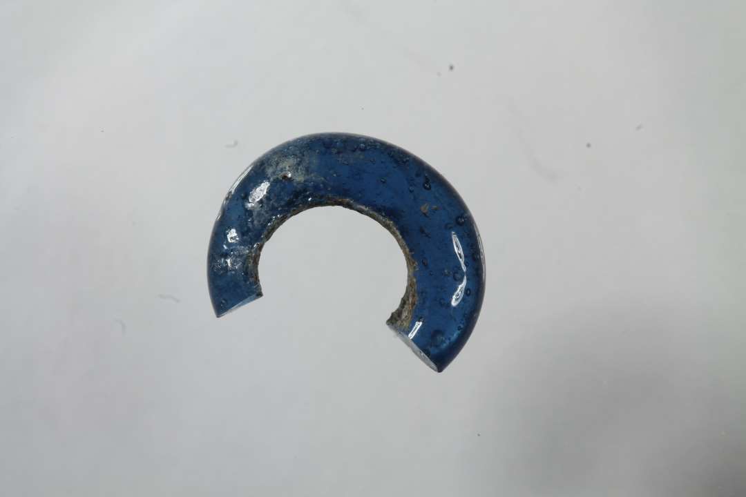 2/3 af ringformet, gennemsigtig, blå glasperle. Diameter: 1,2 cm.