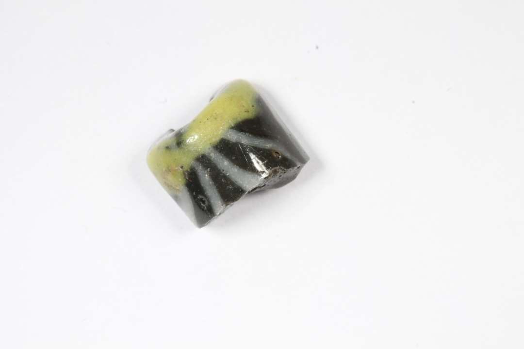 Fragment af cylindrisk, uigennemsigtig sort glasperle med gul og hvid dekoration. Diameter: 0,8 cm.