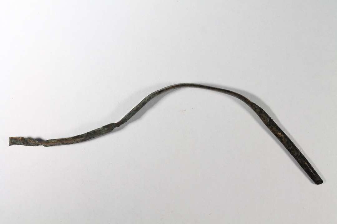 Bronzestang, der for en stor del er hamret flad på den ene side, og svagt buet på den anden. Længde: 9,7 cm., bredde: 0,7 cm.