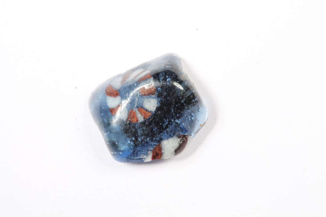 Fragment af polyhydrid, polyedrisk, gennemsigtig blå glasperle med rødt og hvidt. Største mål: 0,8 cm.