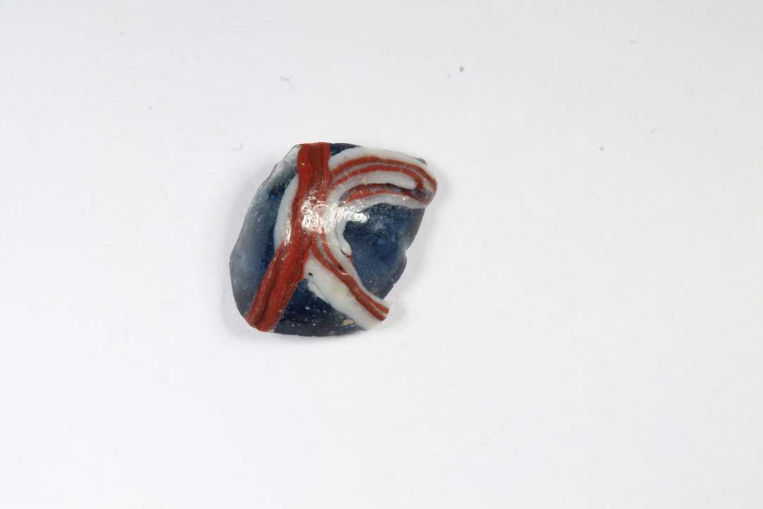 Fragment af cylindrisk ribeperle. Gennemsigtig blå med hvidt og rødt. Største mål: ca 1 cm.