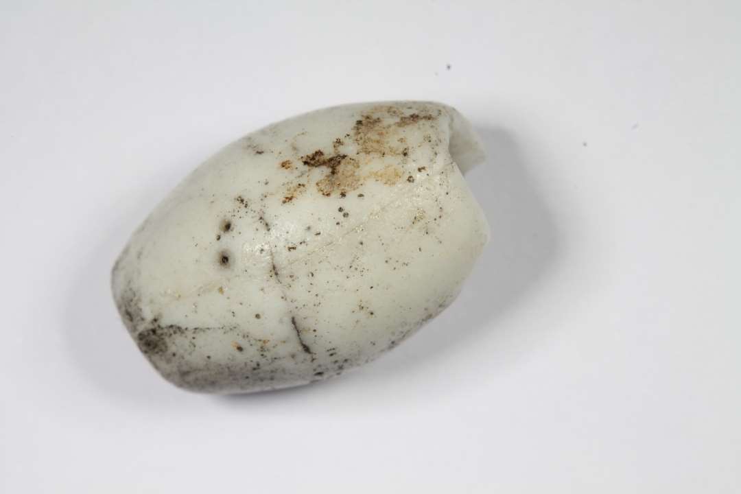 Fragmenter af tøndeformet, uigennemsigtig hvidgrå glasperle, limet sammen til een perle, ca 3/4 del bevaret. Længde: 1,6 cm.