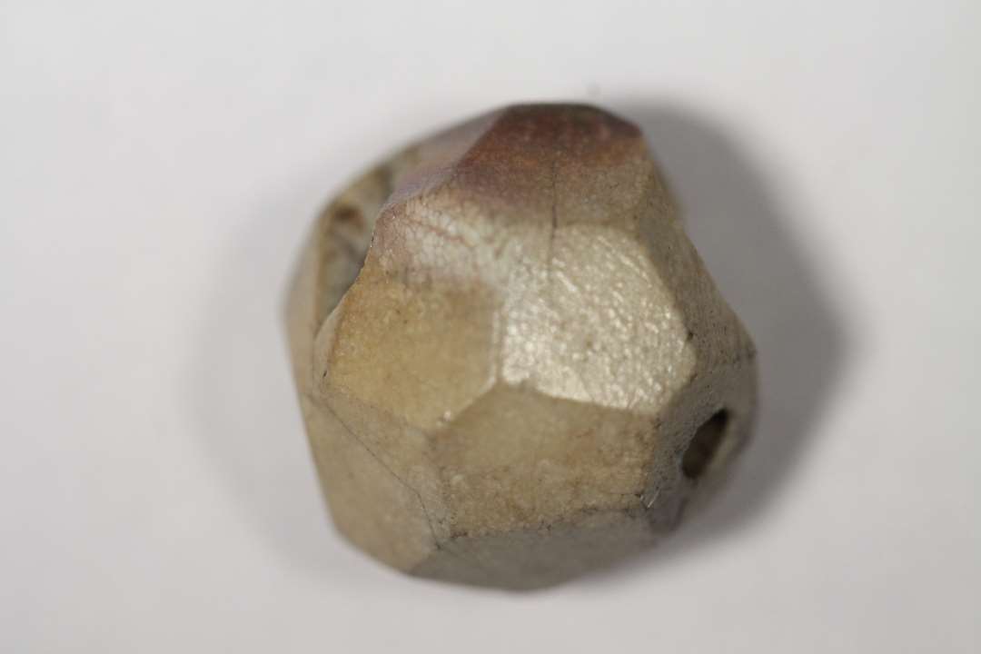 Fragment af perle af stenart. Næsten kugleformet, facetteret med mange små facetter. Lidt mere end halvdelen bevaret. Mål: 1,2x1,2 cm.
Der er antagelig tale om ildskørnet bjergkrystal.