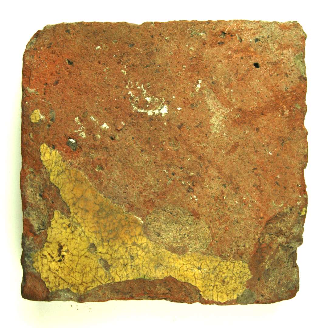Kvadratisk flise af rødbrændt lergods. Flisens overside har oprindelig været dækket af hvid pibelersbegitning, hvorover klar blyglasur. Denne belægning er helt afslidt på nær et parti i flisens ene hjørne. Flisen måler ca. 16,7 - 8 cm. i kvadrat. Tykkelse: 3,3 - 3,6 cm.