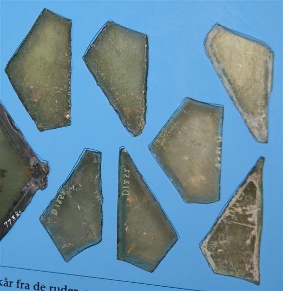 214 fragmenter af klart, grønligt glas, med tildels afknappede kanter fra ruder dels i form af ligebenede, spidsvinklede trekanter, dels i form af skæve spidsvinklede trekanter.