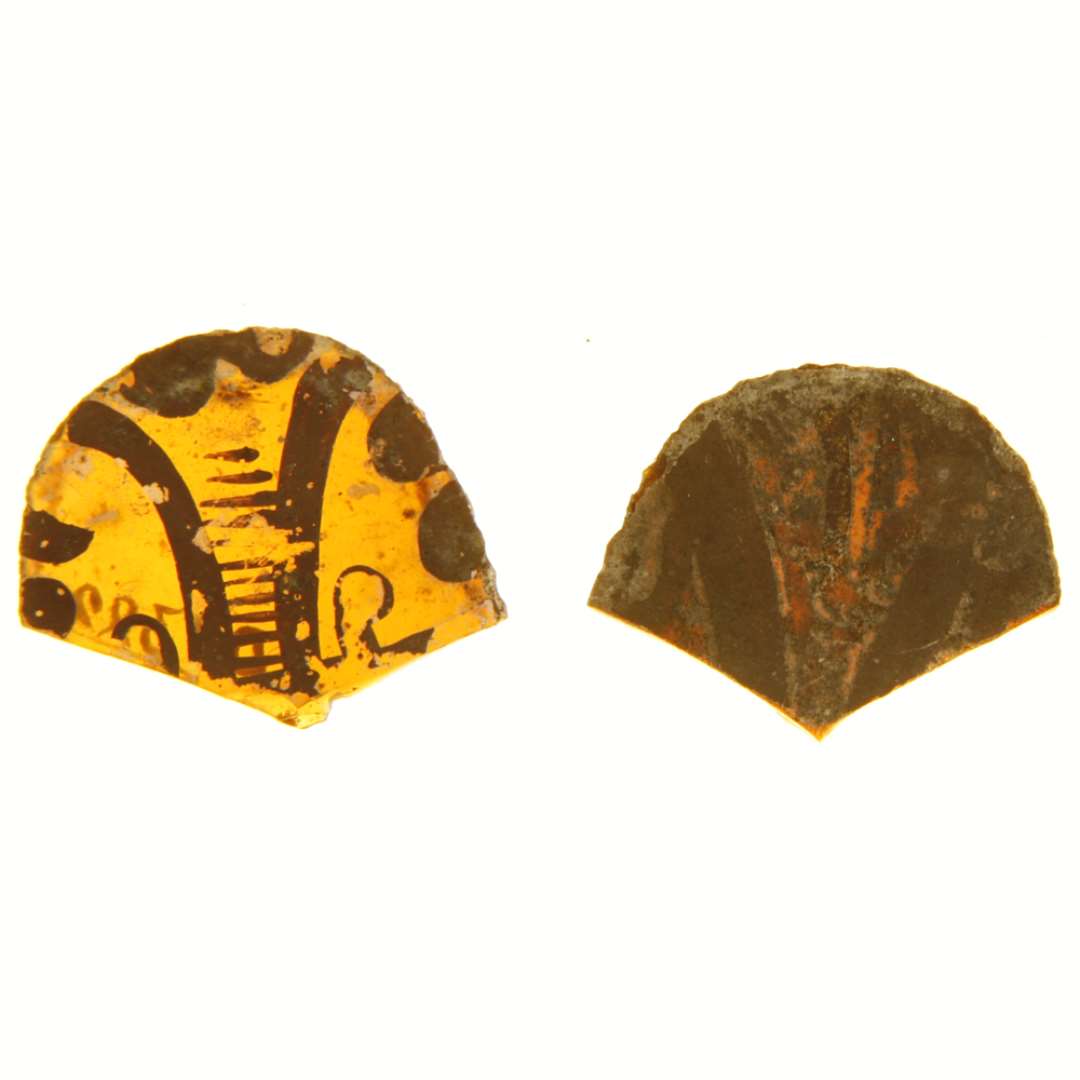 2 fragmenter af ruder af gult glas med afnappede kanter bemalet med stiliserede plantemotiver i brunlige farver. Hidrører antagelig fra ruder af kort, bred tungeform, men kun tungespids partiet er bevaret. Største bredde og længde: ca. 3 x 2,5 cm. og 3,1 x 2,7 cm.