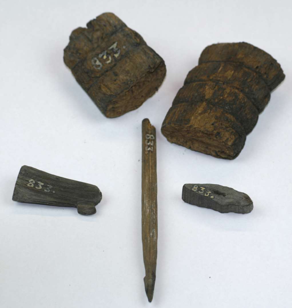 Fragmenter af fire genstande af træ.  1 stk.: Pølsepind. Længde: 11,5 cm. 2 stk.: uspec. træfragmenter. Største længder: 3,5 og 4,3 cm. 1 stk.: Fragment af håndtag i to dele. Med omløbende, indskårne bælter. samlet længde: 12 cm.