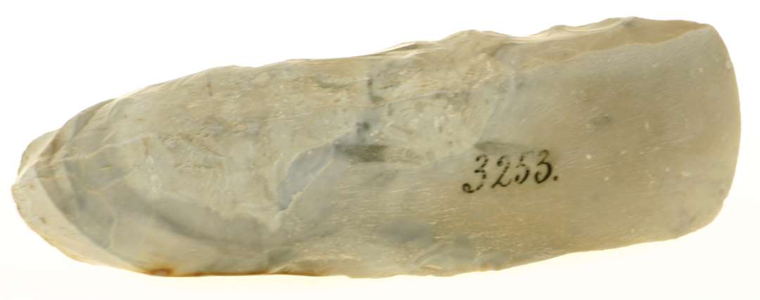 En tyknakket Flintøxe, 14½ Cm. lang og 4½ Cm. bred. Skjæv Nakke med Rest af Skorpe. Kun Bredsidernes Egparti slebent.